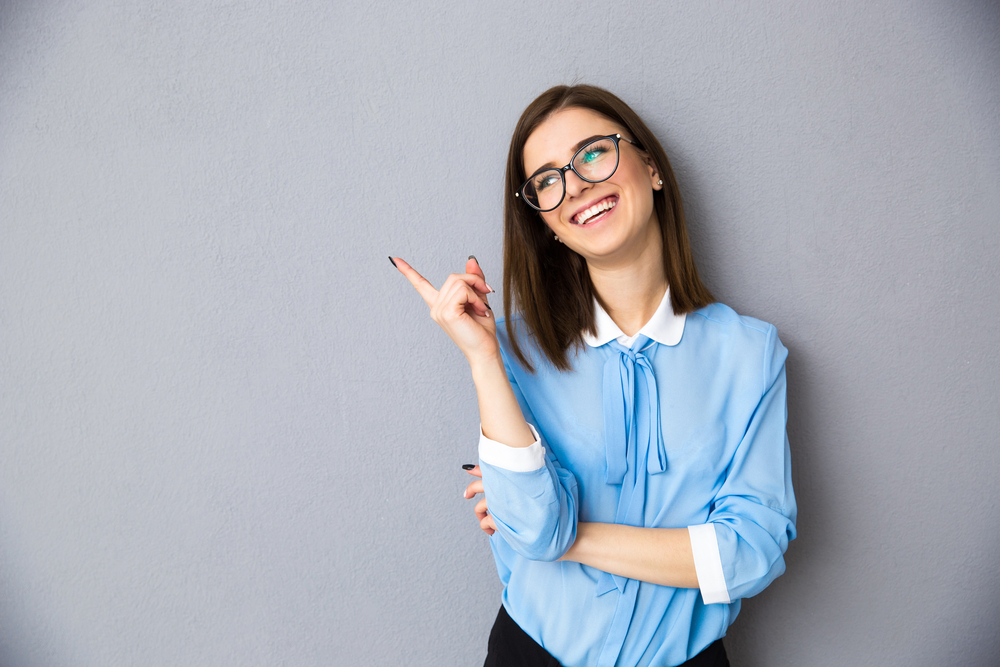 Cheerful businesswoman pointing away over gray background. Wearing in blue shirt and glasses. Looking away