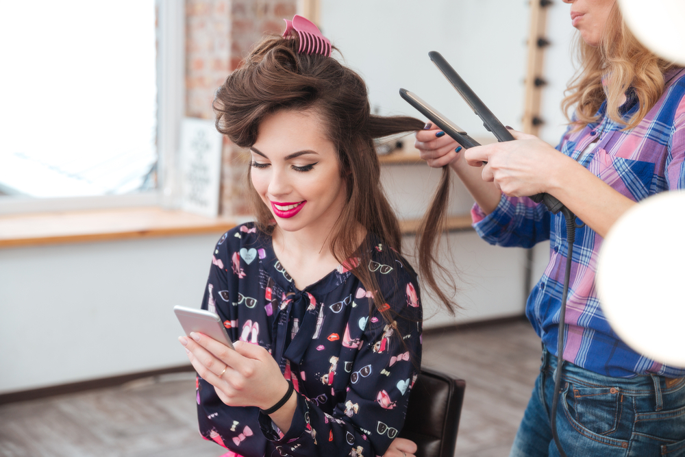 Female hairdresser applying hair straightener for long hair of smiling young woman using smartphone in dressing room